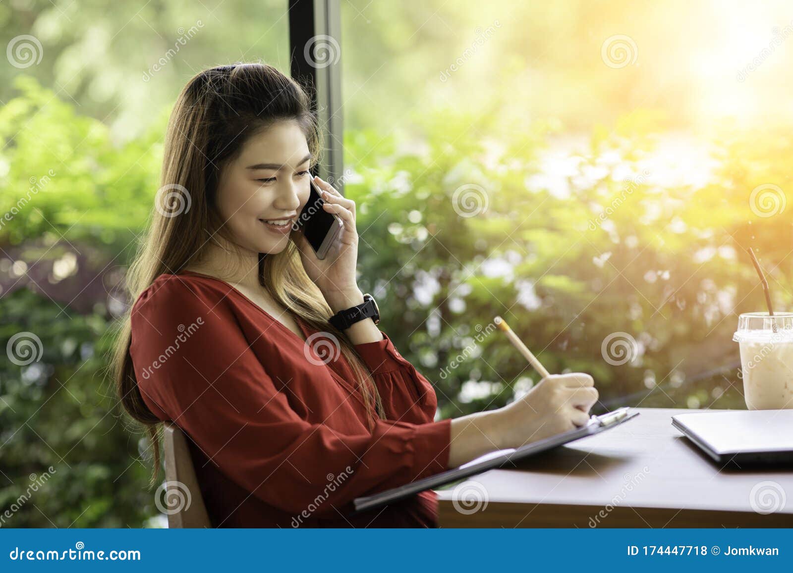 attractive asian woman wearing red dress working with laptop smile and talking with customer in smartphone