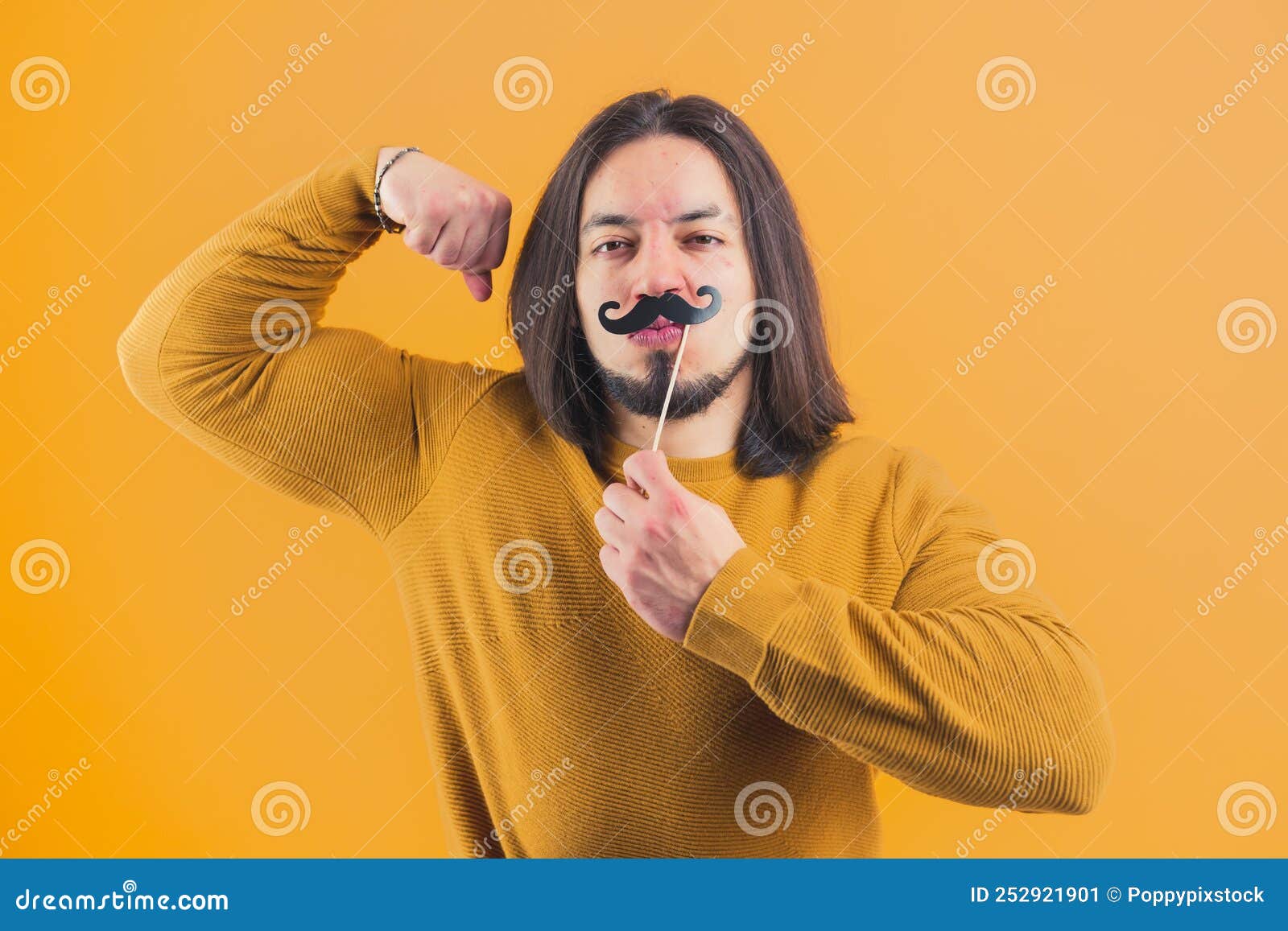 attractive bearded handsome hispanic boy hanging paper moustache and tighting muscles on a yellow background in a studio