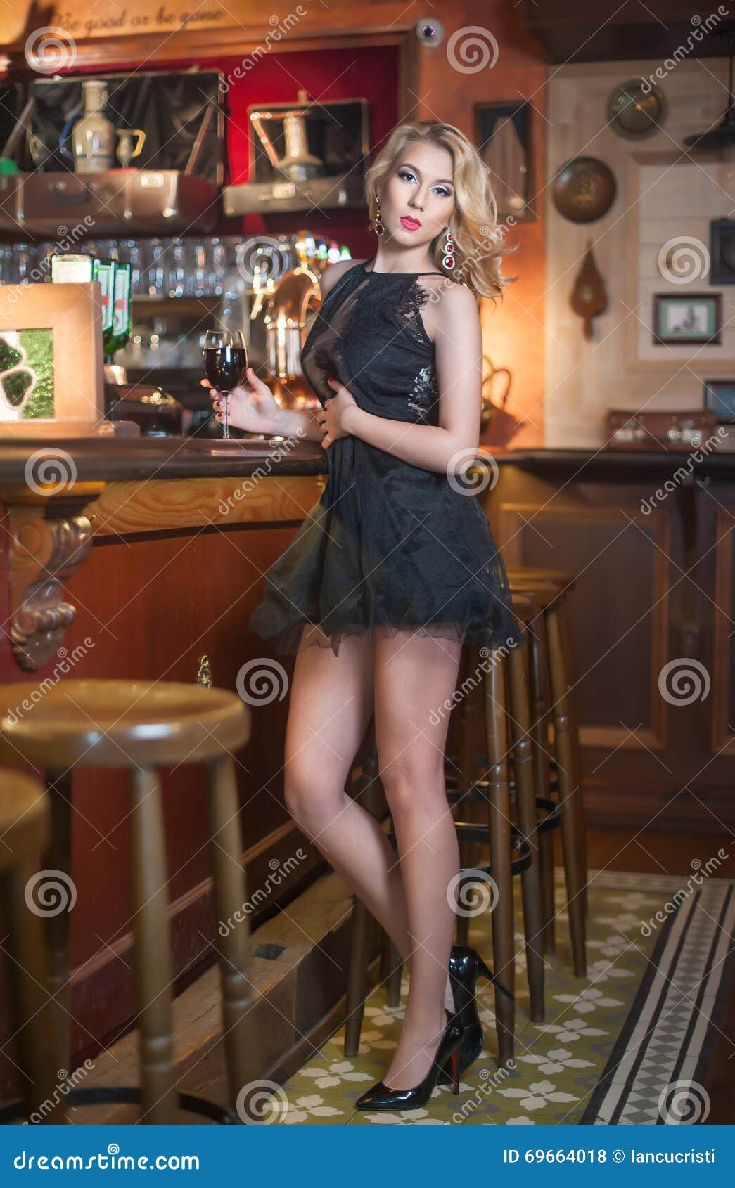Attractive Blonde Woman With Curly Hair In Elegant Short Lace Dress Standing Near Bar Stool