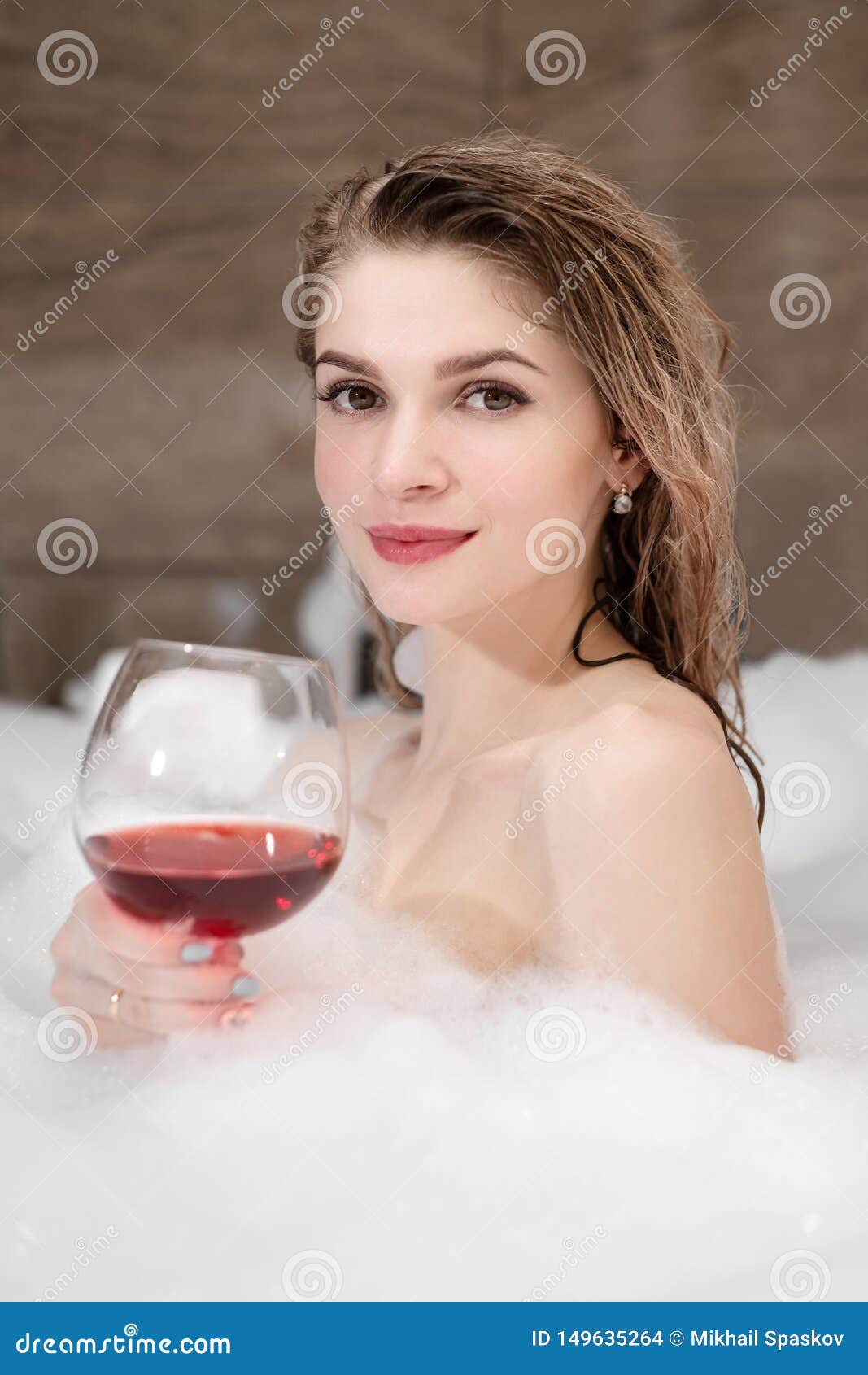 Attractive Blonde Woman Lying In Bathtub In Foam In Hotel Drinking Red Wine From A Large Glass