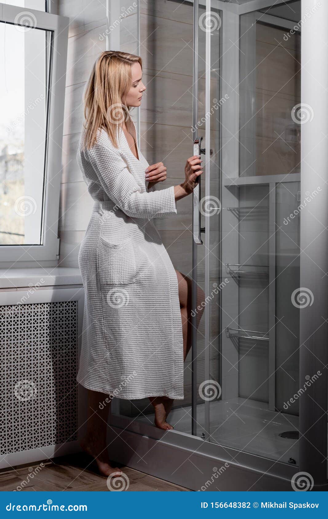 Attractive Middle Age Blond Woman In White Bathrobe In Bathroom He Enters The Glass Shower And