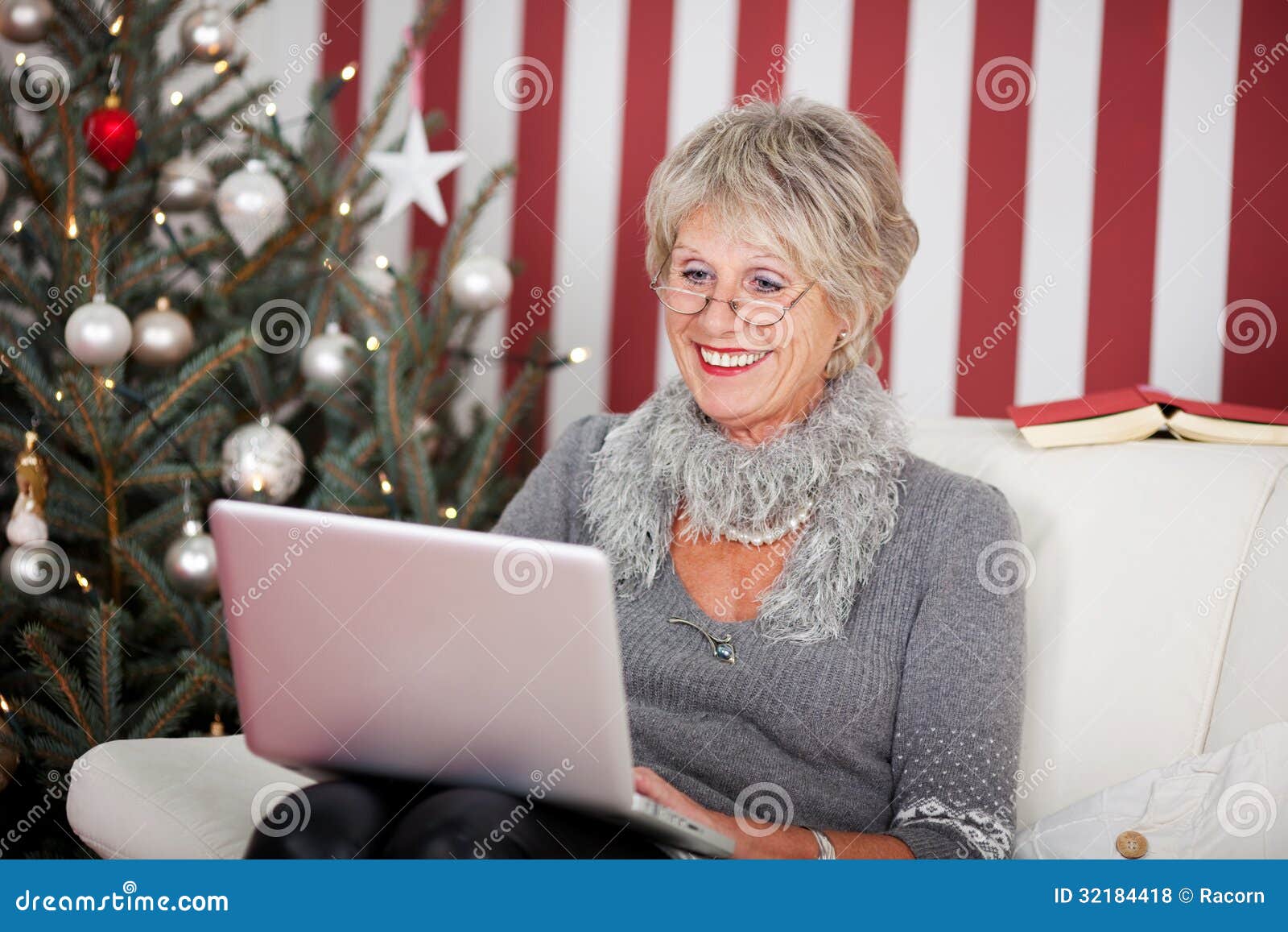 Attractive senior using a laptop. Attractive glamorous senior woman sitting in her living room in front of a decorated Christmas tree using a laptop, red and white themed