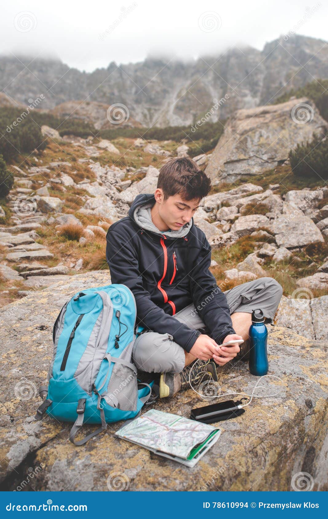 Aufladung eines Handys auf einem Gebirgspfad. Junge, der auf einem Felsen stillsteht und einen Handy auflädt