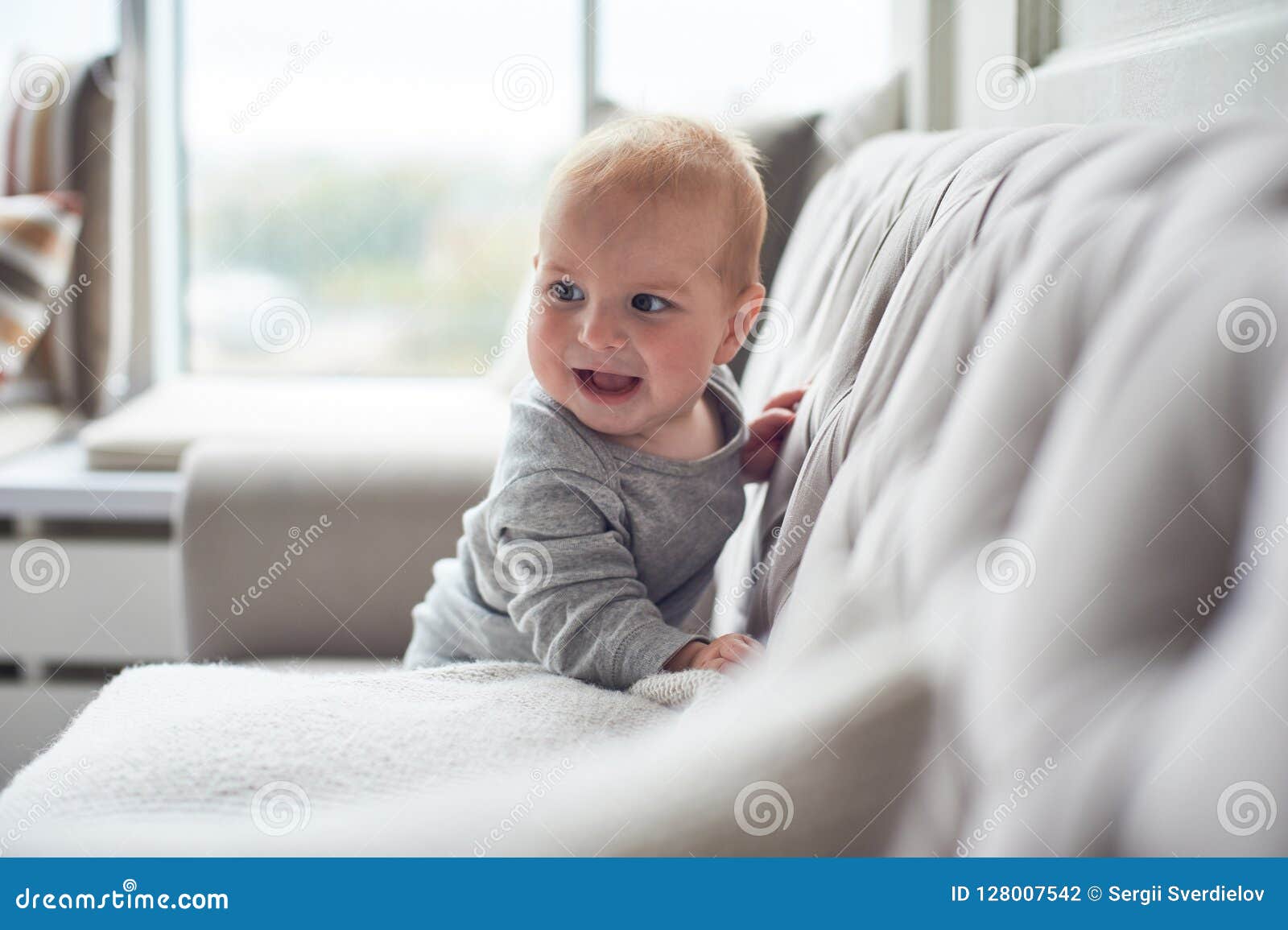 Baby Boy Crawling and Climbing on Sofa Against Big Window Stock Photo