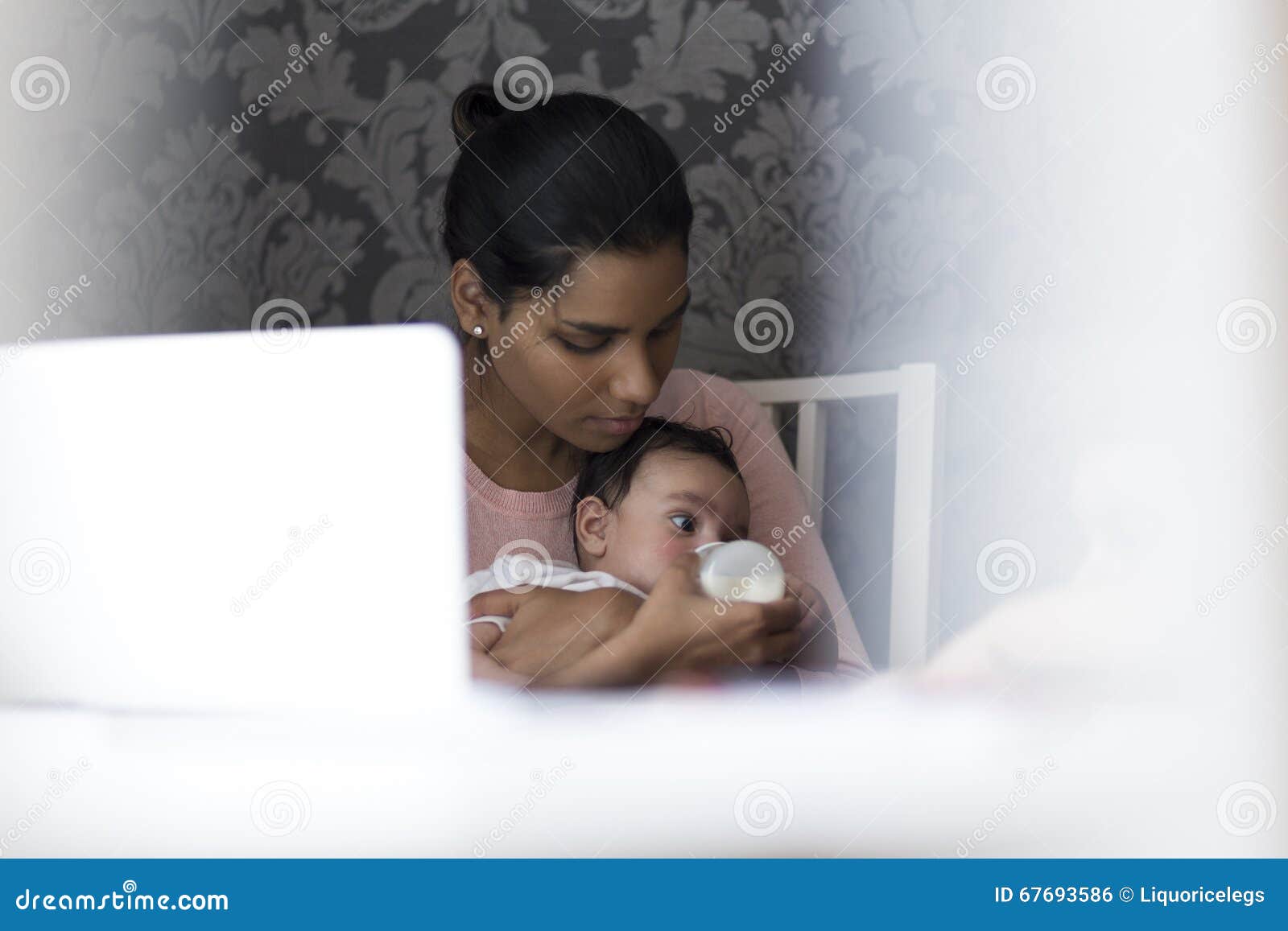 Baby feeding time. Young mother feeding her baby son a bottle of milk whilst sitting at her computer.