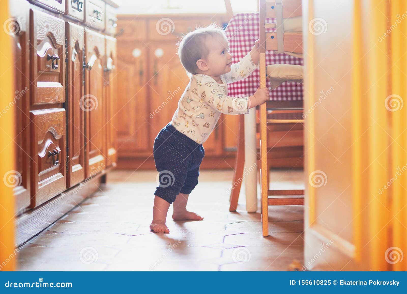 Baby Girl Standing On The Floor In The Kitchen And Holding On To