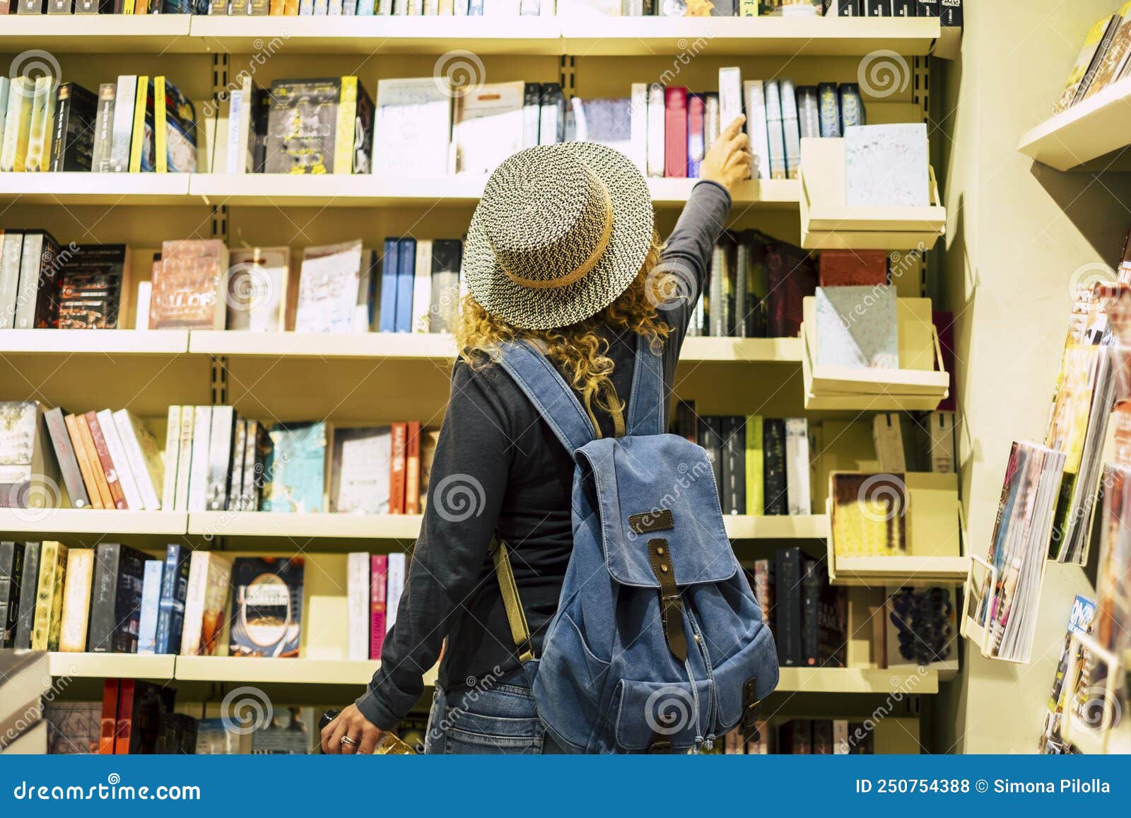 back view of woman with hat and backpack choosing a book to buy inside a library or newspaper store. people prepare to travel.