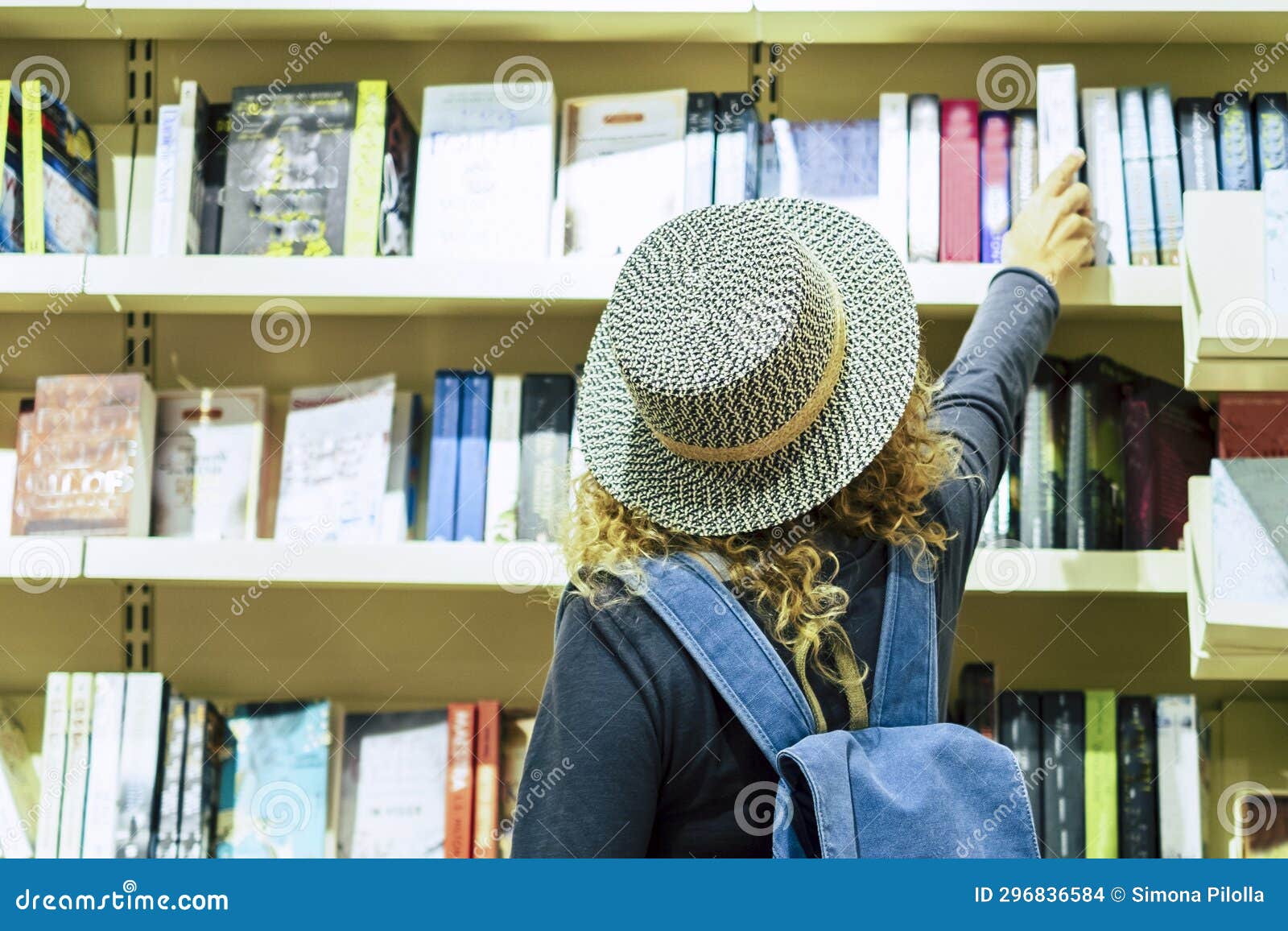 back view of woman with hat and backpack choosing a book to buy inside a library or newspaper store. people prepare to travel.