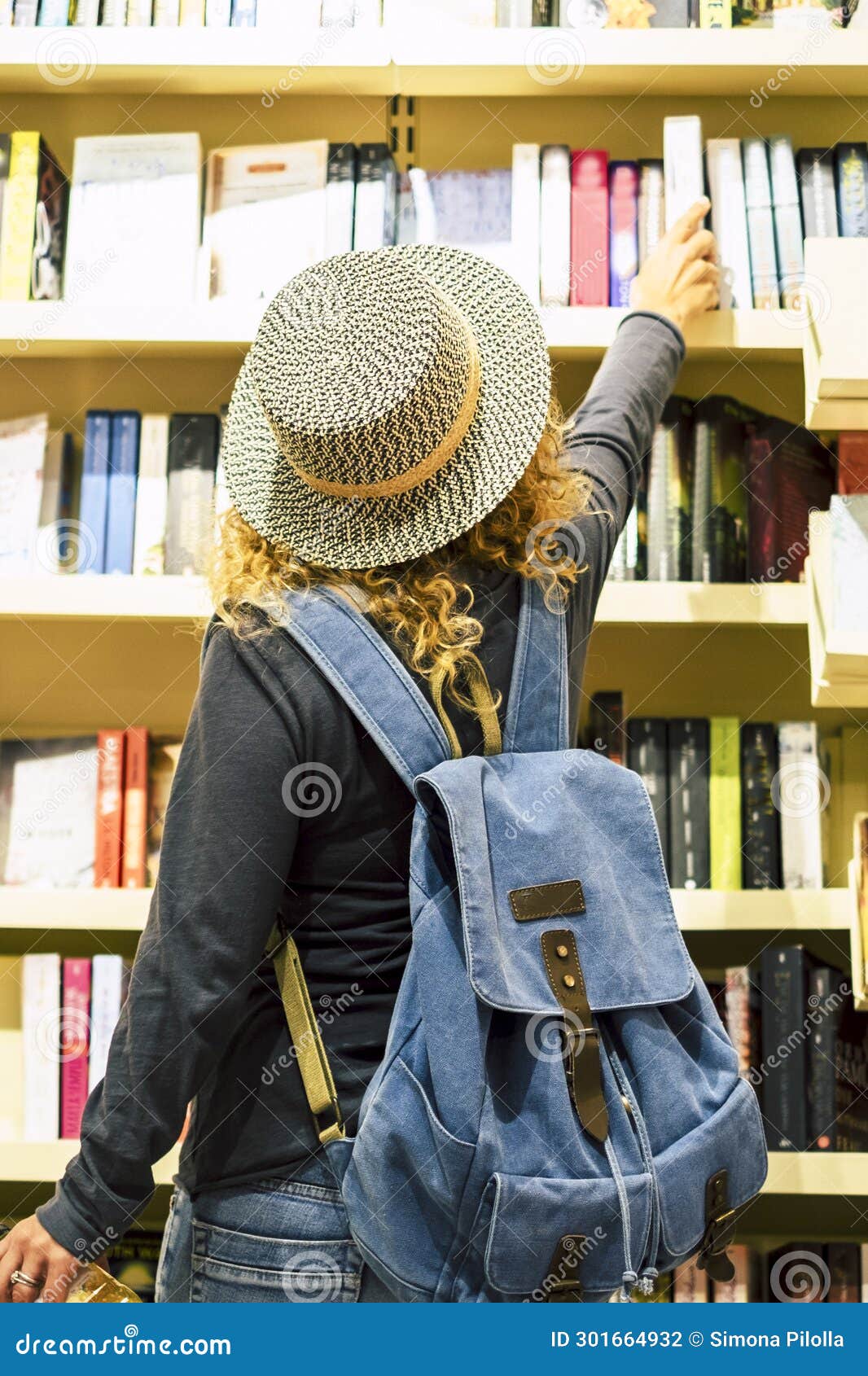 back view of woman with hat and backpack choosing a book to buy inside a library or newspaper store. people prepare to travel.