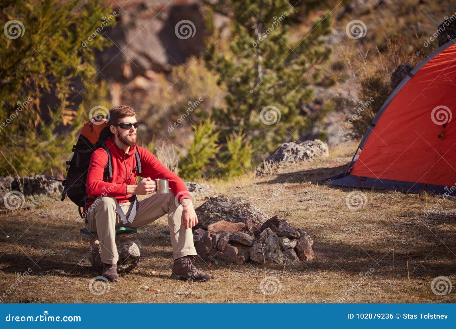 backpacker having rest near orange tent with cup in hand.