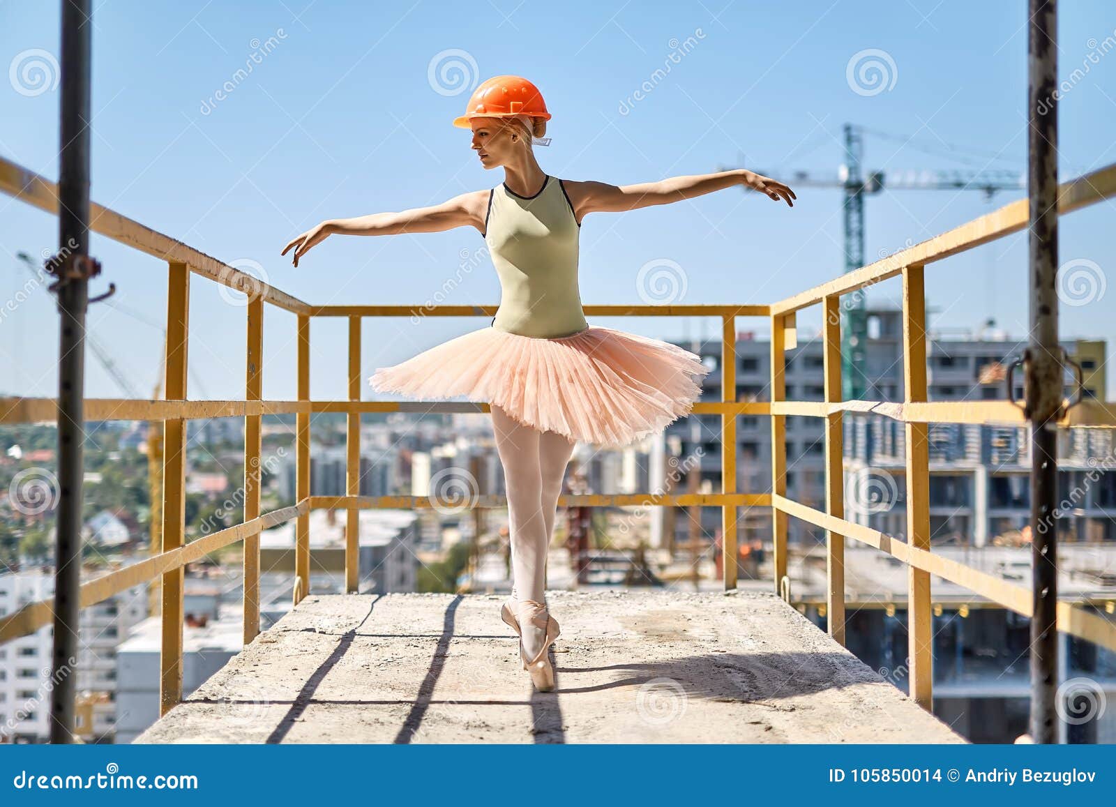 Ballerina som poserar på den konkreta balkongen. Den glade ballerina står på tårna på den konkreta balkongen av den oavslutade byggnaden på cityscapebakgrunden Hon bär den gröna bodyen, persikaballerinakjolen, damasker, pointes och konstruktionshjälmen