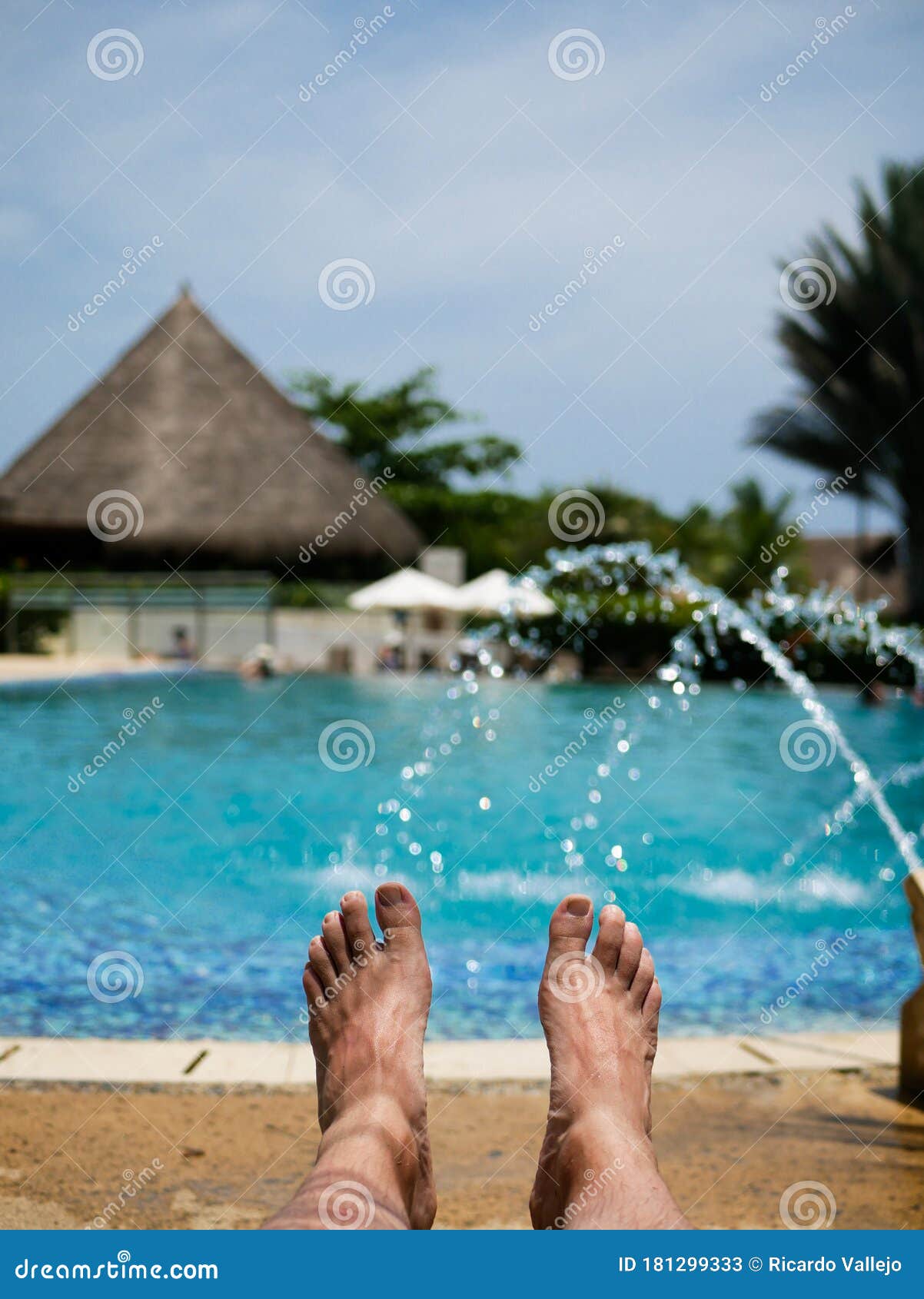 barefoot feet of a man taking a sun bath in a pool with fountains nd a hut on the background