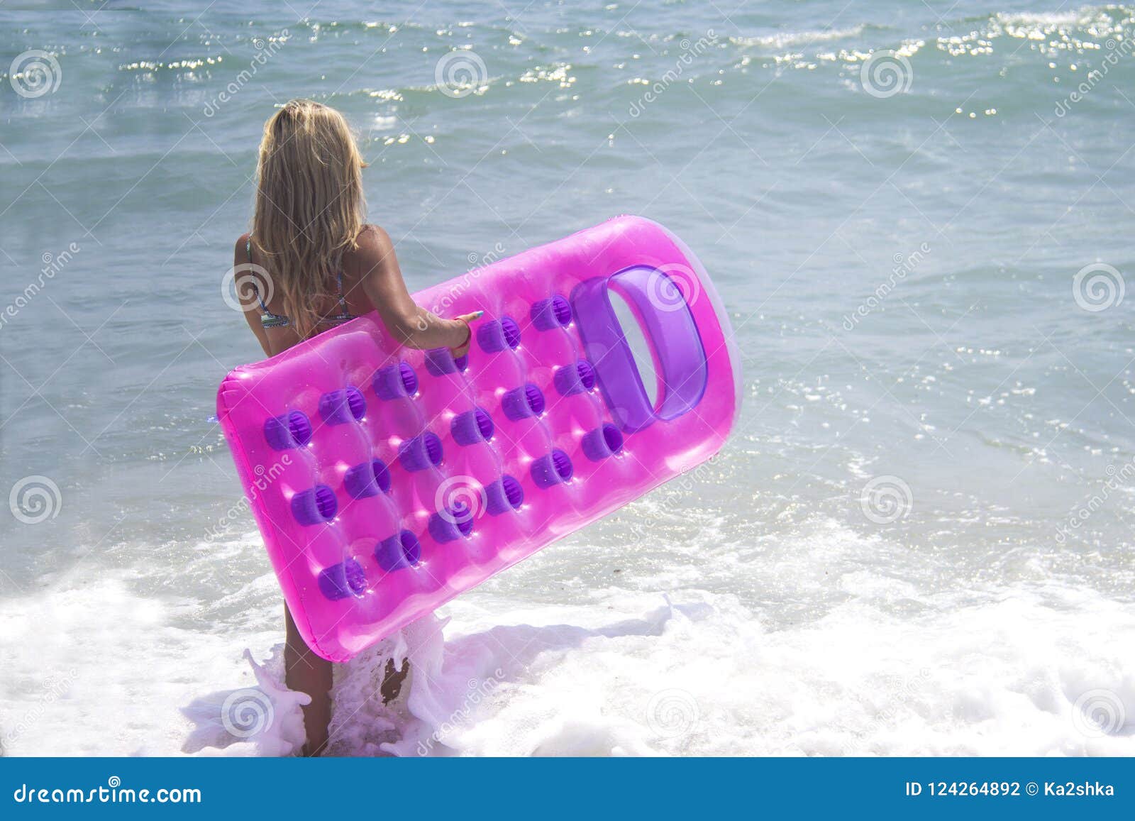 Barfüßigmädchen im blauen Bikini in dem Meer Nehmen Sie das große Mädchen in der Badebekleidung gehend in das Meer mit rosa aufblasbarem Poolfloss ab