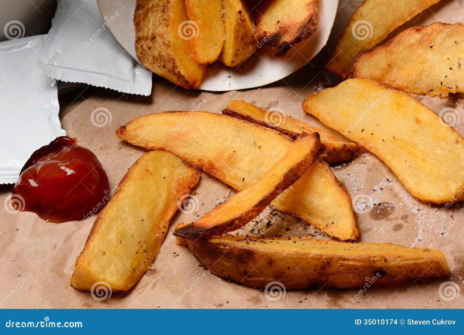 Batatas fritas com ketchup no saco de Brown. O close up de certas batatas fritas derramou em um saco marrom. Dollop e pacotes da ketchup com sal e pimenta. Formato horizontal que enche o quadro.