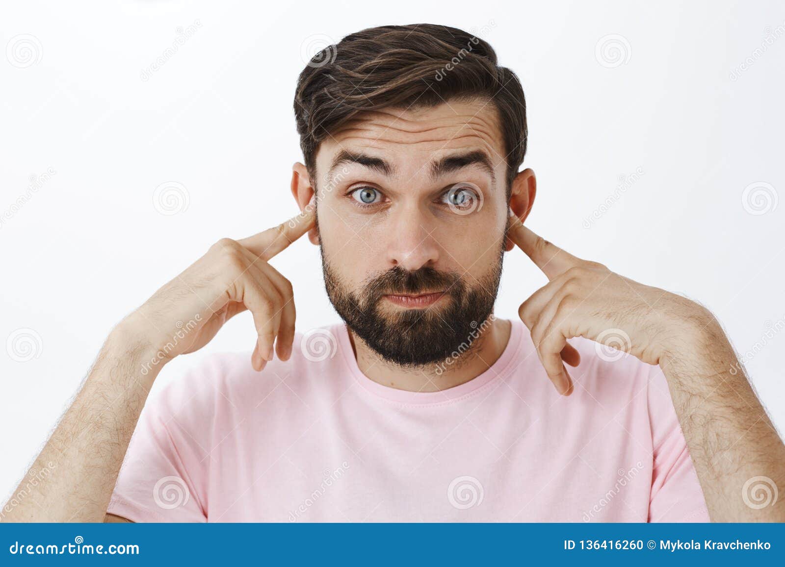 bearded caucasian man close ears as standing in loud crowded place, holding index fingers in earholes pouting and