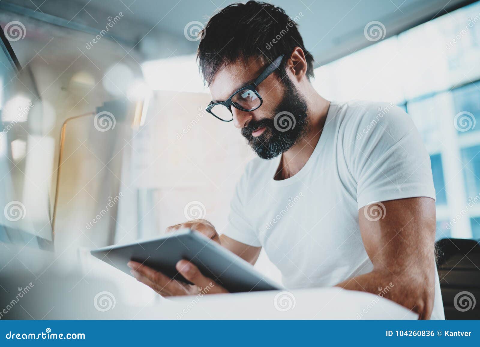 bearded man in white tshirt wearing eye glasses and using portable electronic pro tablet computer at modern lightful