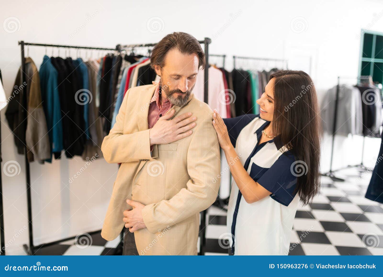 Assistance avec le montage Beau styliste de attirance rayonnant aidant un mâle attirant adulte aux cheveux foncés barbu beau tout en essayant sur un nouveau blazer