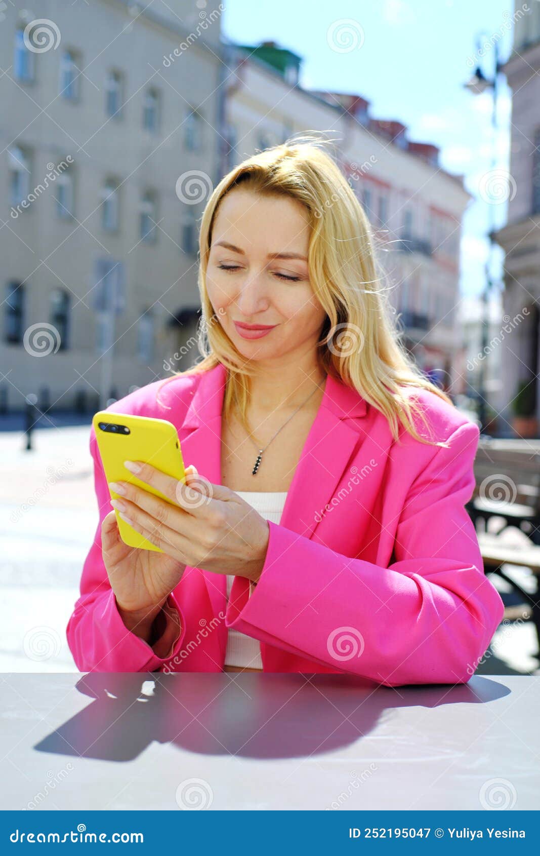 a beautiful adult blonde woman in a bright fuchsia-colored jacket sits at a table in a cafe and looks at the screen of a