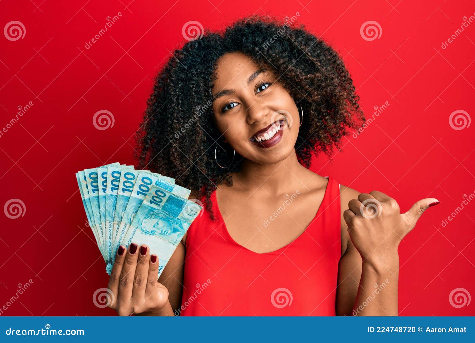 beautiful african american woman with afro hair holding 100 brazilian real banknotes pointing thumb up to the side smiling happy