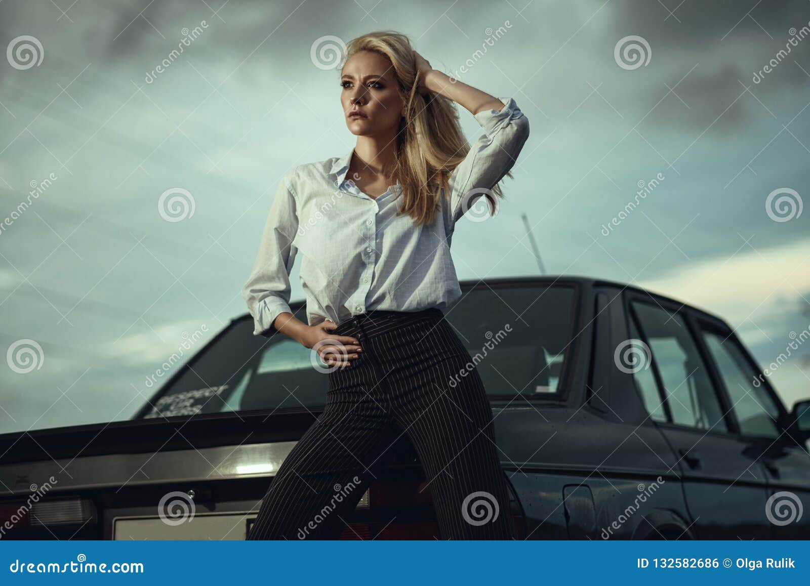 beautiful blond lady in black striped high waisted pants and oversized white blouse standing at her old car looking away