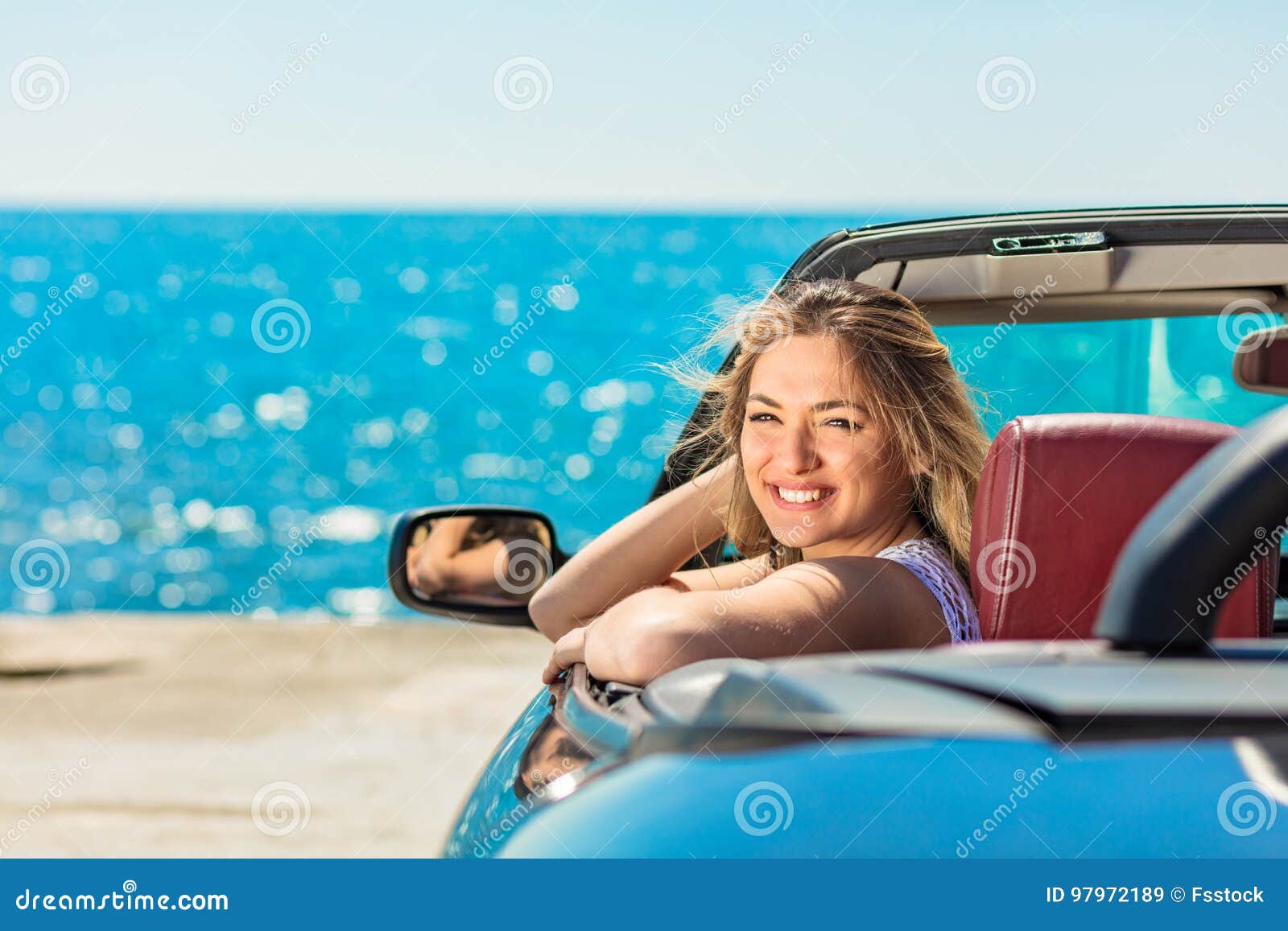 beautiful blond smiling young woman in convertible top automobile looking sideways while parked near ocean waterfront