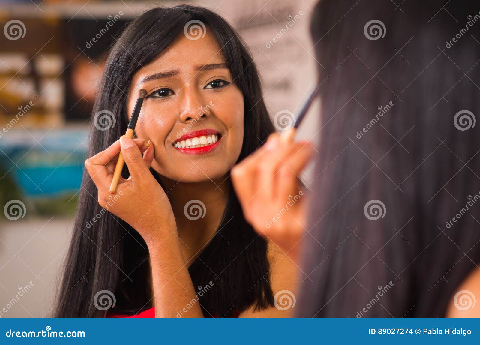 beautiful brunette applying make up while smiling happily, seen from behind and face reflecting in mirror