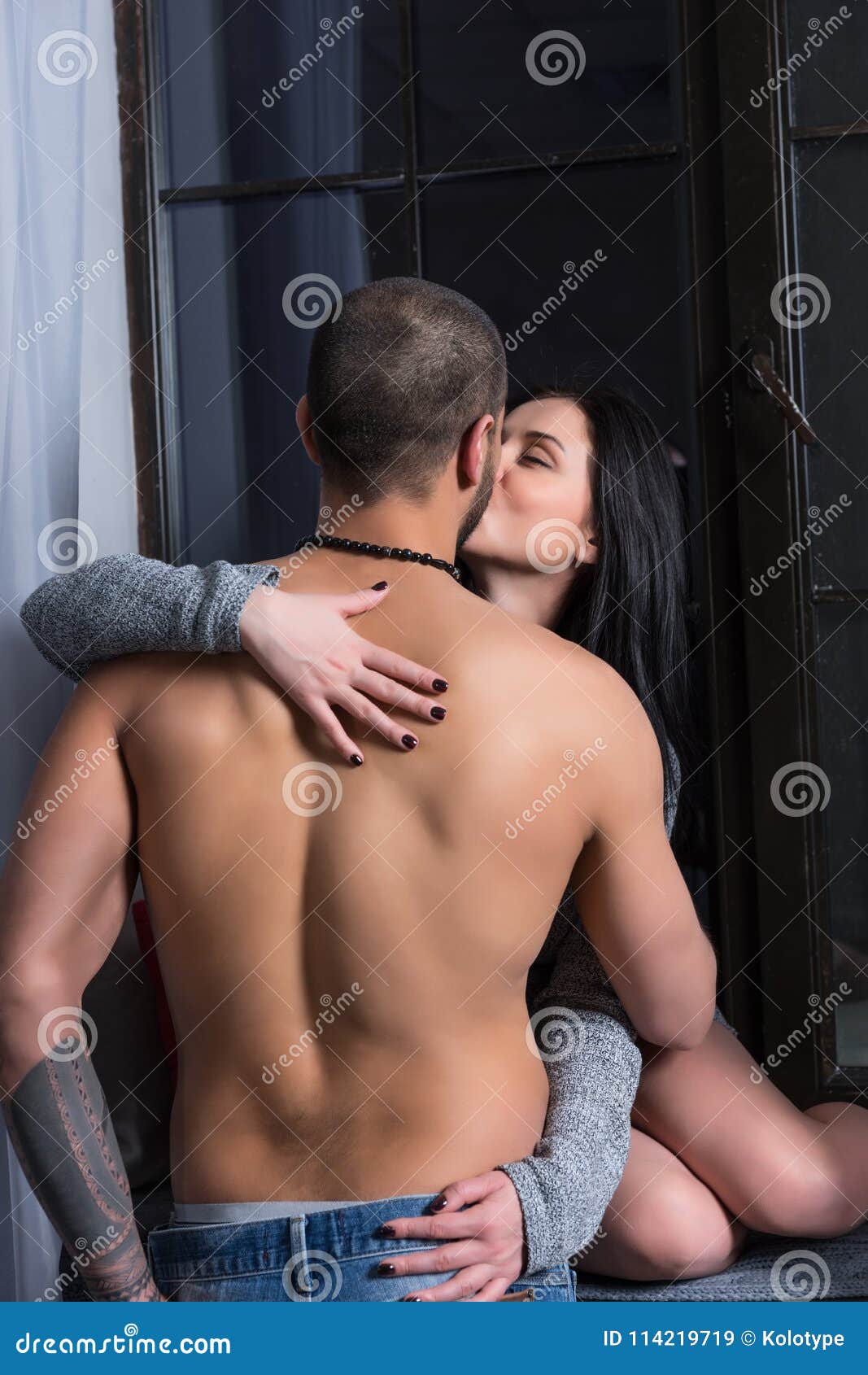 Beautiful Brunette Woman in a Knitted Dress Sits on the Windowsill and Kissing Her Husband with Bare Chest Stock Image