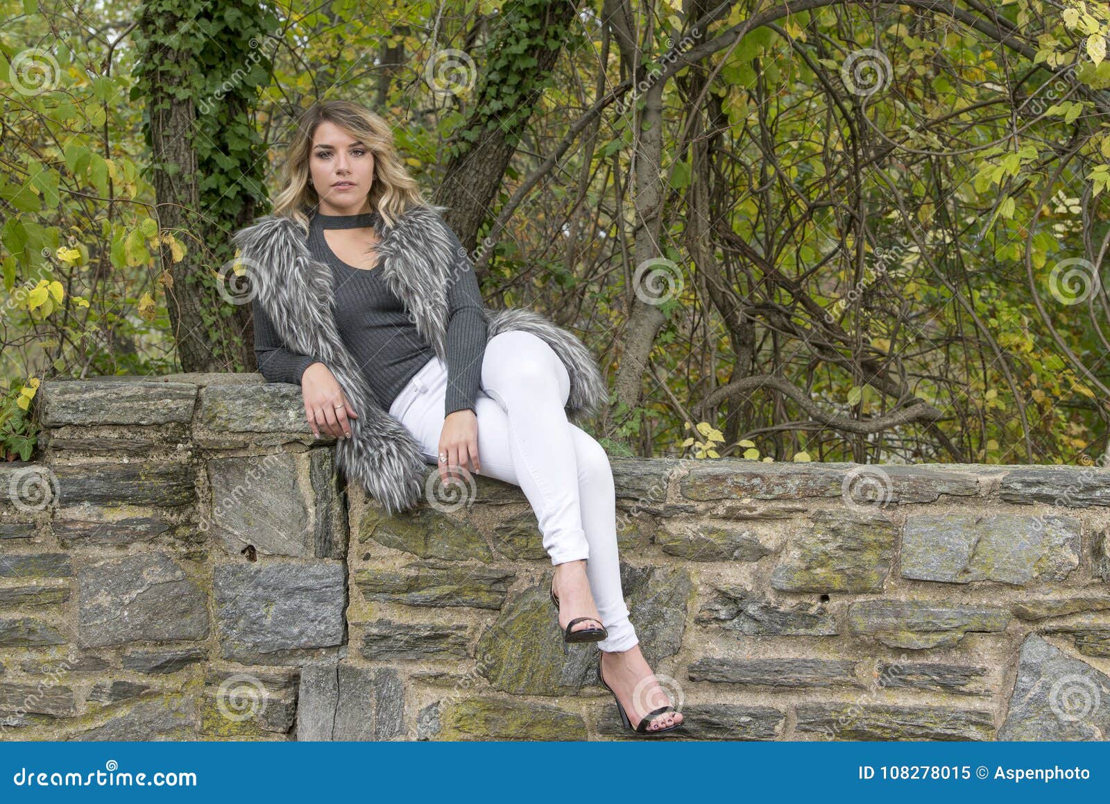 beautiful caucasian female model poses wearing faux fur vest white pants park autumn sitting stone wall outdoor 108278015