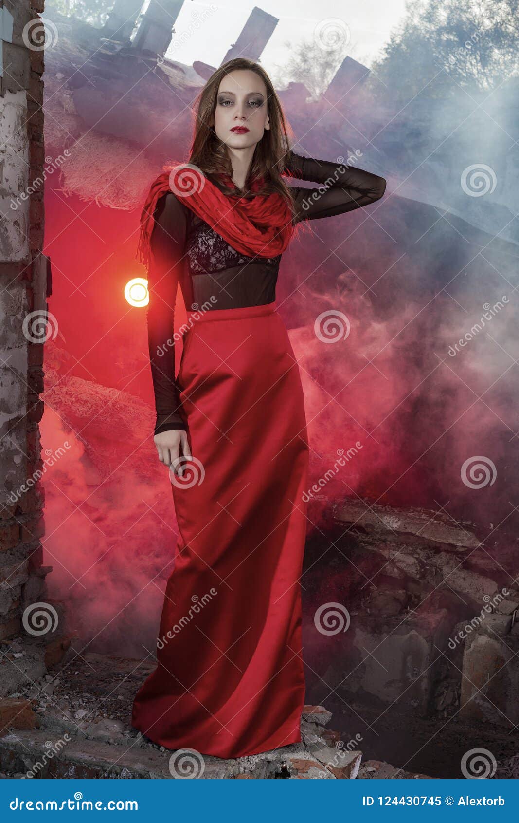 Beautiful Fashion Model Girl Wearing A Red Long Skirt And A Red Shawl Poses  At The Collapsed Building, Among The Ruins Of Which Rises Red Smoke.  Conceptual, Fashionable, Modern And Advertising Design.