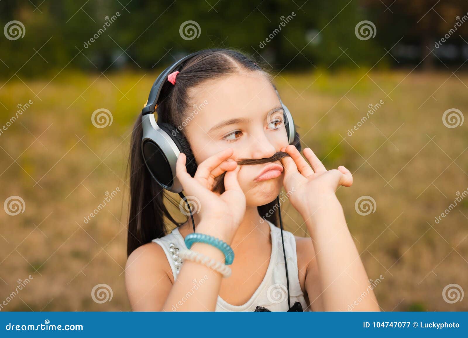 Beautiful girl making funny faces in nature. Hipster serious girl having fun shows moustache hair over field. child listening music with headphones