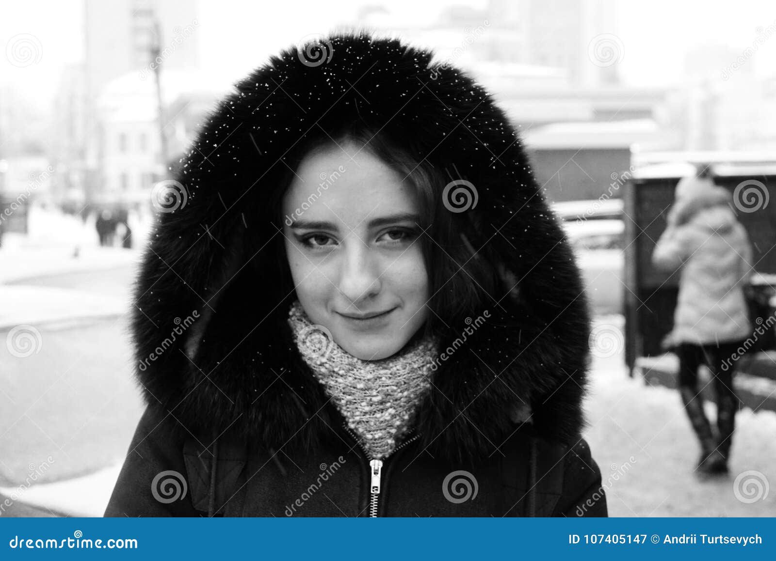 Beautiful girl on the winter streets of a city. A beautiful girl is walking down a street. It is winter time and it is snowing. She is wearing a coat with a hood and a scarf, looking toward you. She has long brunette hair. Snowflakes on her hood.