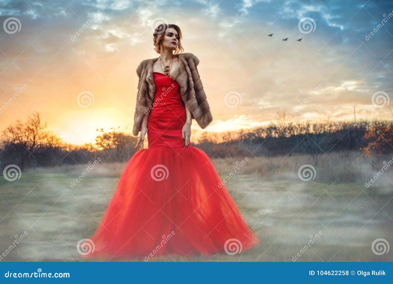 beautiful glam model with updo hair wearing posh red fishtail dress and luxurious mink vest standing in the misty field at sunset