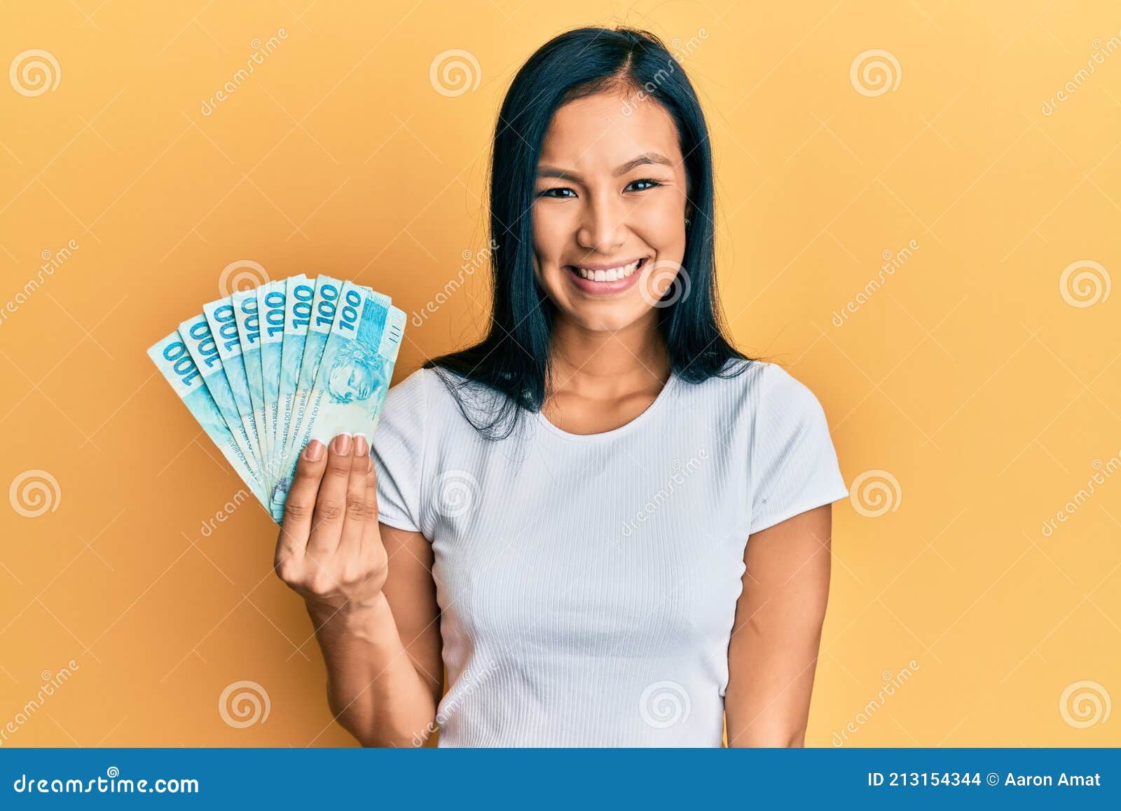beautiful hispanic woman holding 100 brazilian real banknotes looking positive and happy standing and smiling with a confident