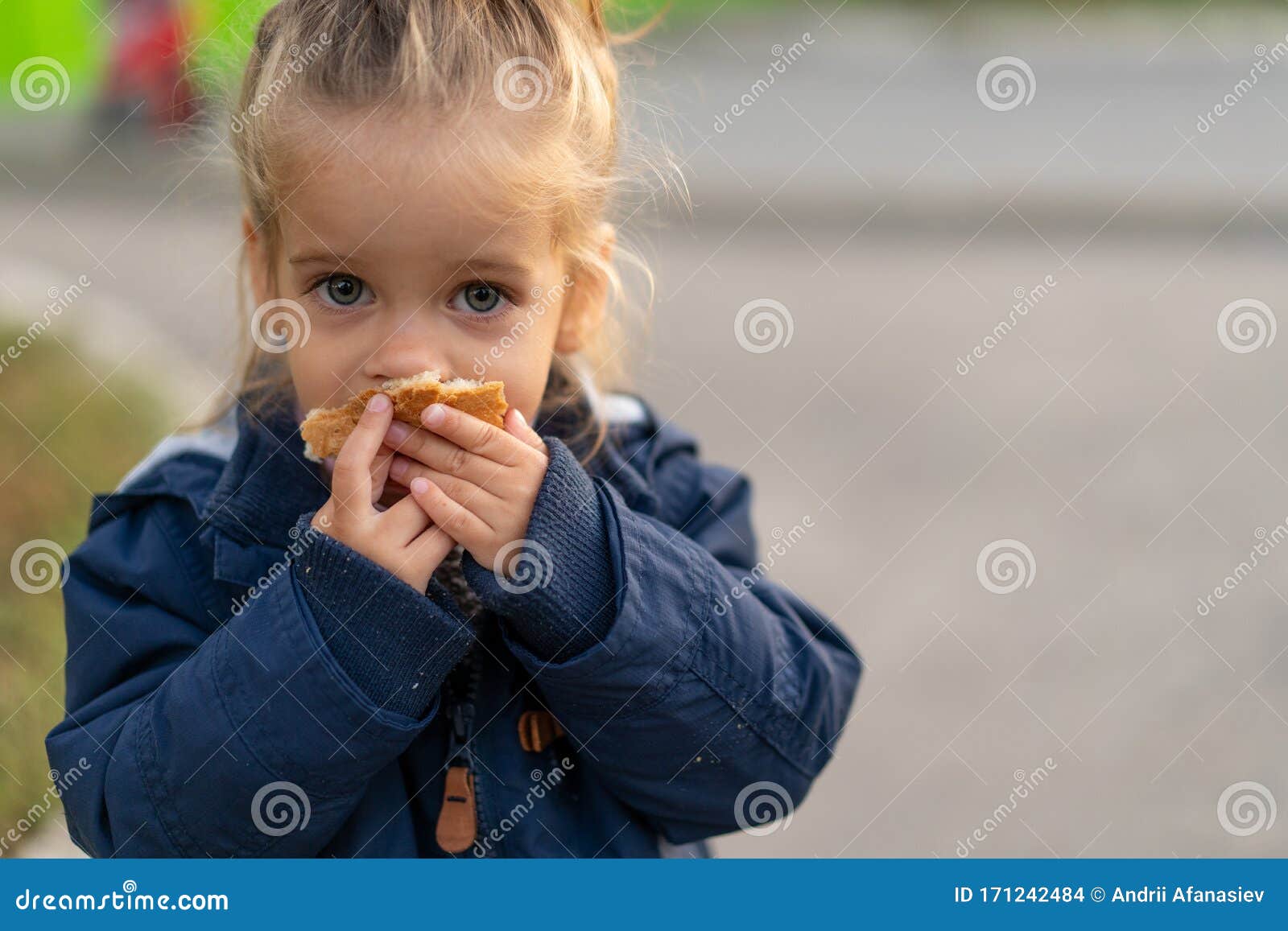 a beautiful little caucasian girl with blond hair and eating bread eagerly with her hands looks at the camera with sad eyes