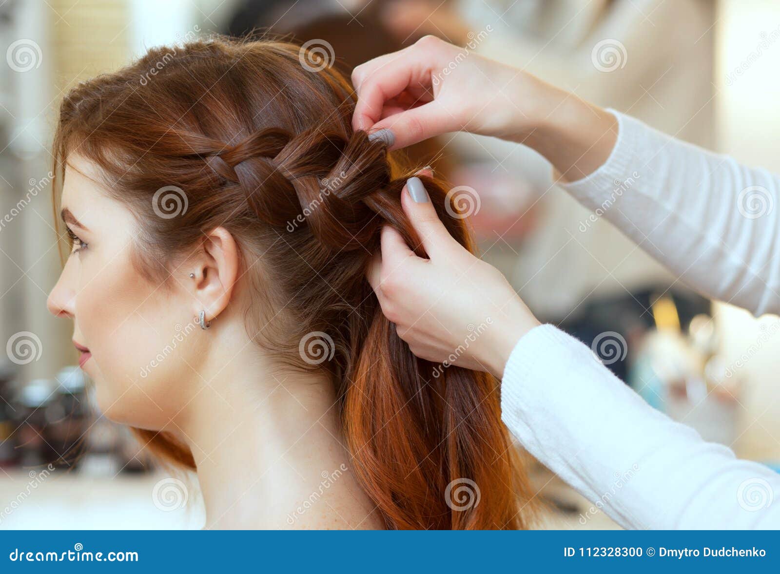 beautiful, red-haired girl with long hair, hairdresser weaves a french braid, in a beauty salon