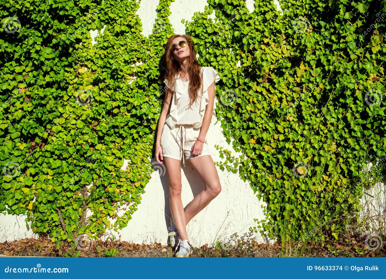 beautiful relaxed woman in white romper, keds and trendy round mirrored sunglasses standing at the wall entwined with ivy