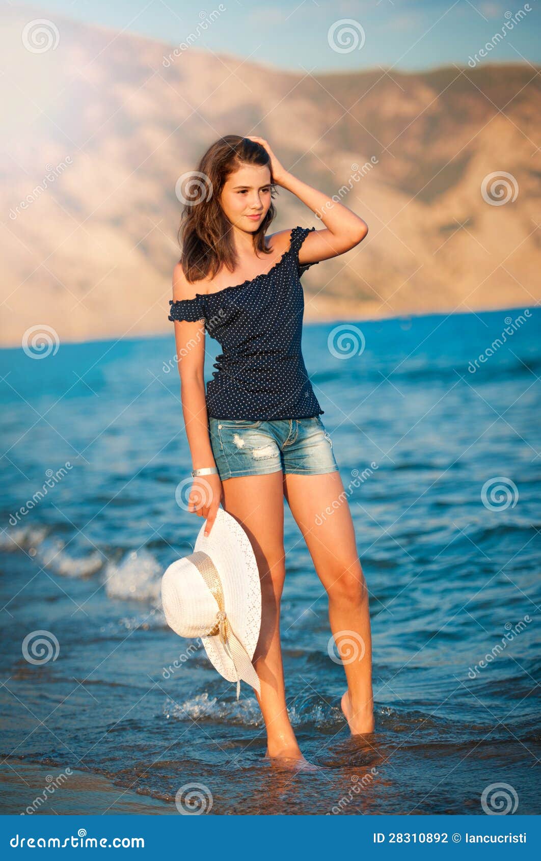 Beautiful Teen Girl Goes On Coast Of Ocean With Straw Hat In Hands
