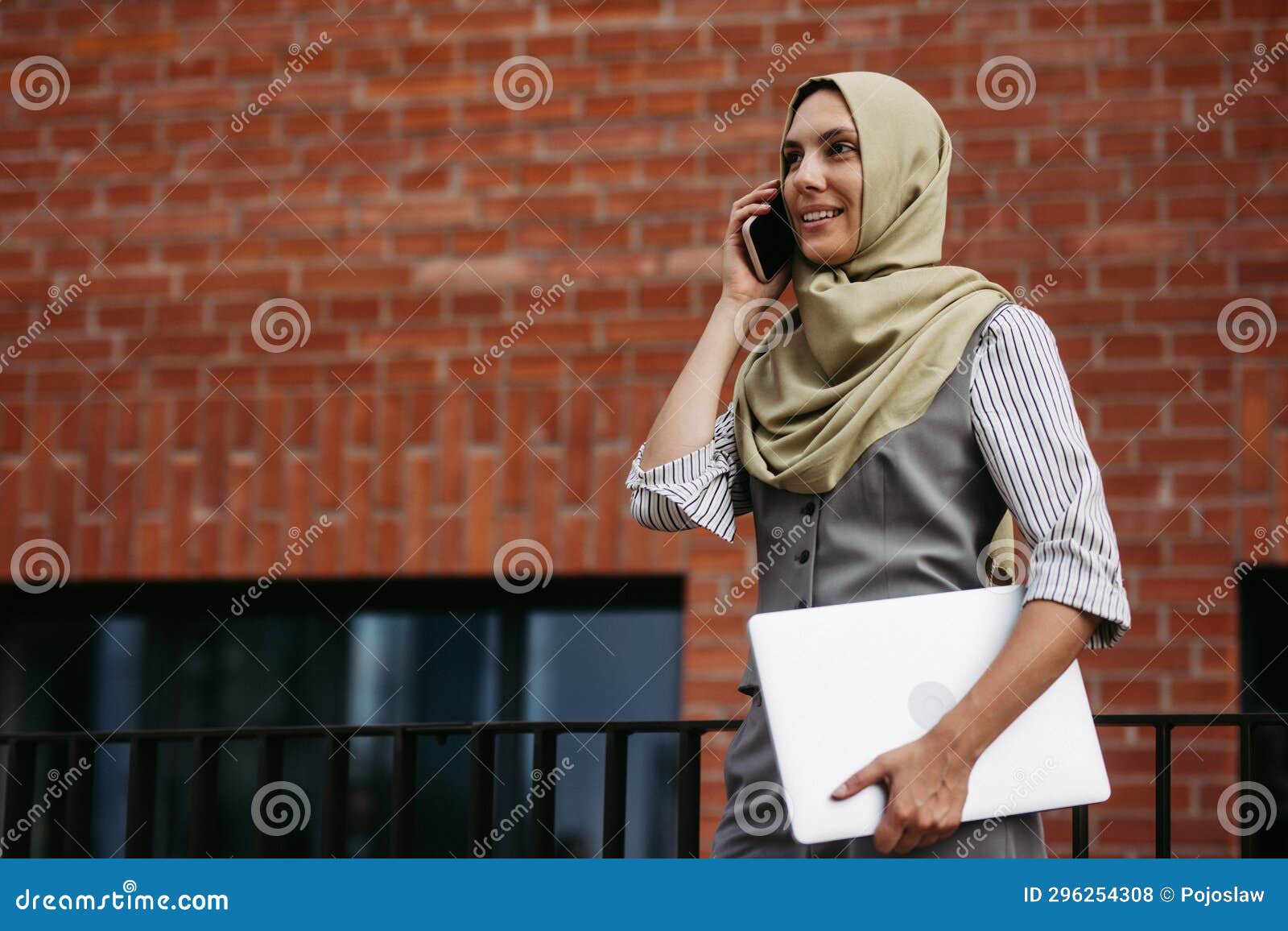 beautiful woman in hijab standing on city street. muslim businesswoman with laptop making call. iran, afganistan female
