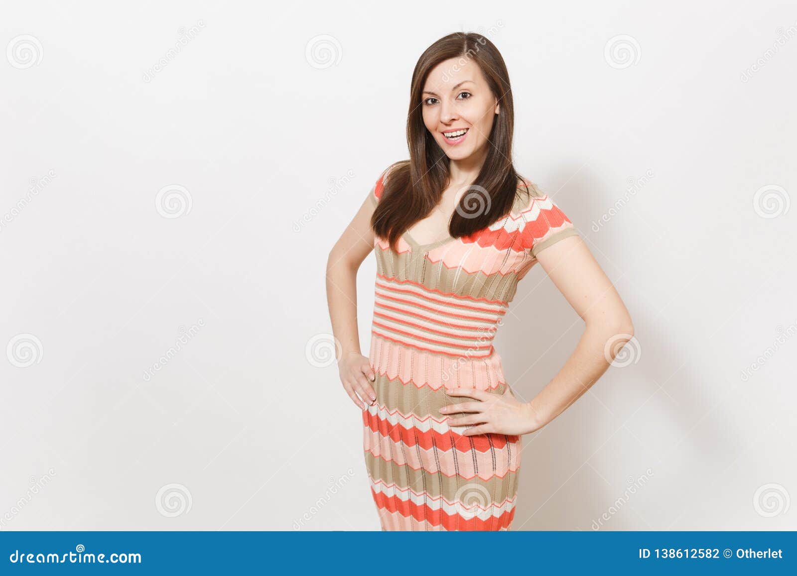 Beautiful young brunette girl in light beige and pink patterned dress rejoices, smiling and holding hands at waist in studio on white background. Concept of good mood.