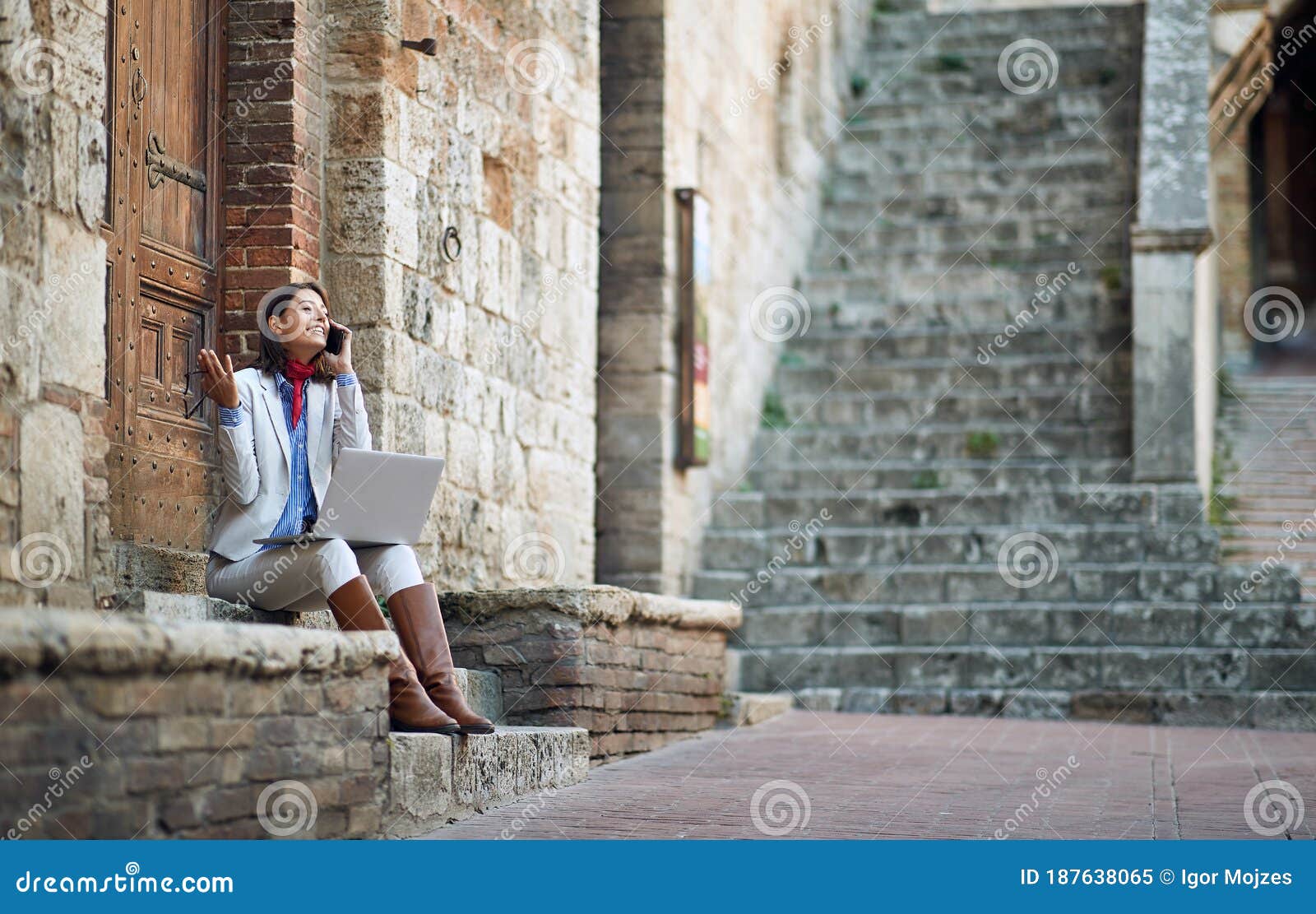 young female talking on her cell phone with laptop, sitting outdoor on stairways in italy