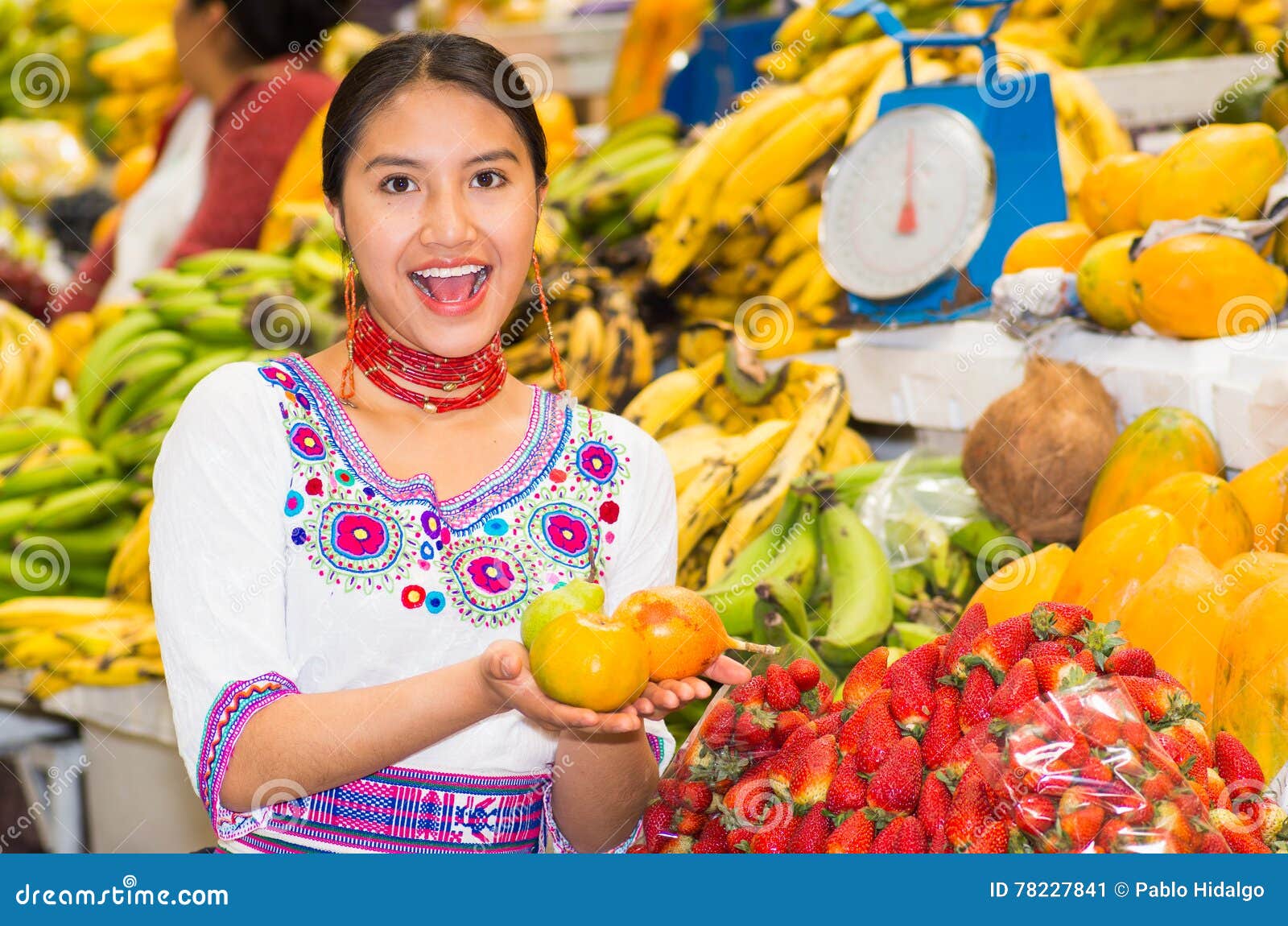 Beautiful young hispanic woman wearing andean traditional blouse posing for camera holding granadillas and smiling, inside fruit market, colorful healthy food selection in background.