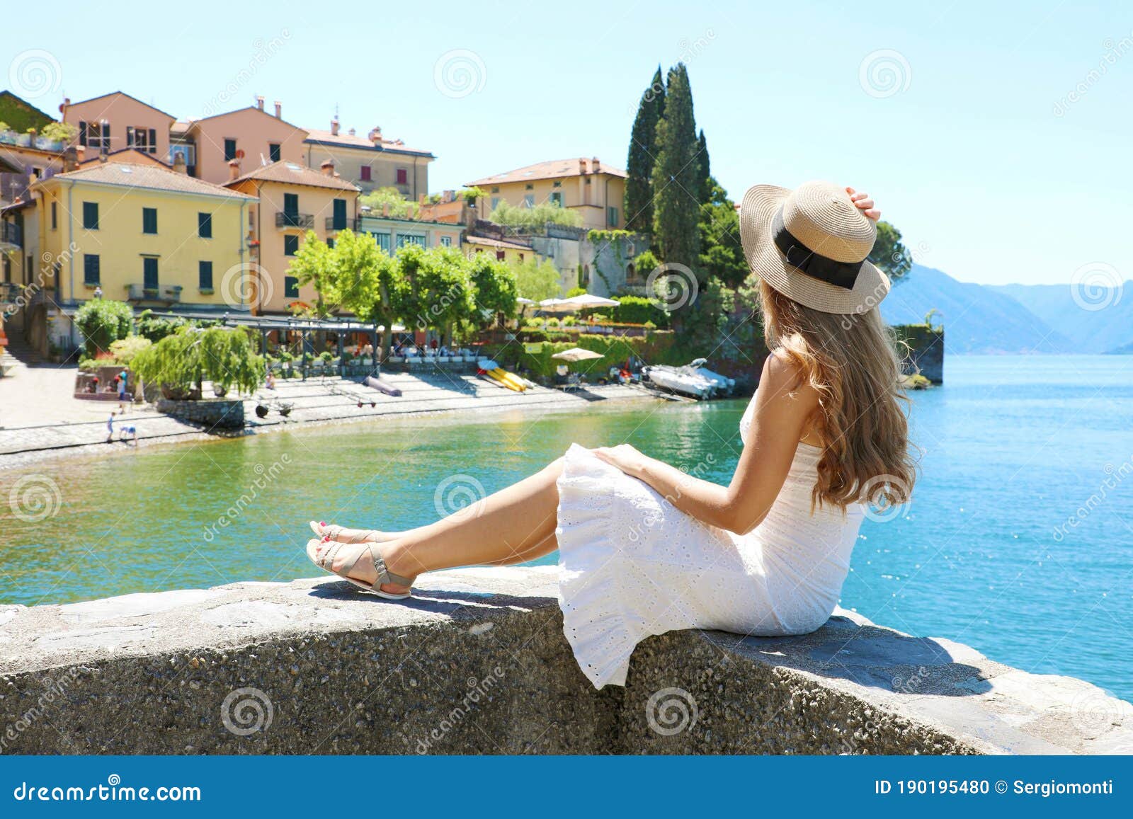 beautiful young woman with hat sitting on wall looking at varenna town on lake como, italy