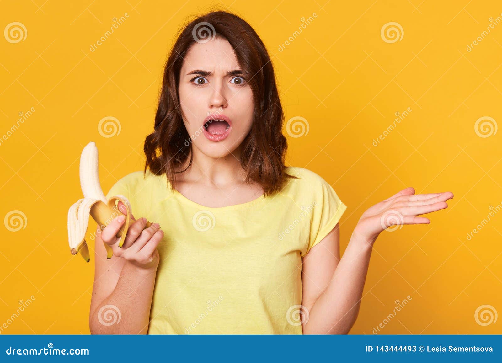 beautiful young woman holding fresh banana, takes hands to side, has uncomprehending facial expresion, standing with open mouth
