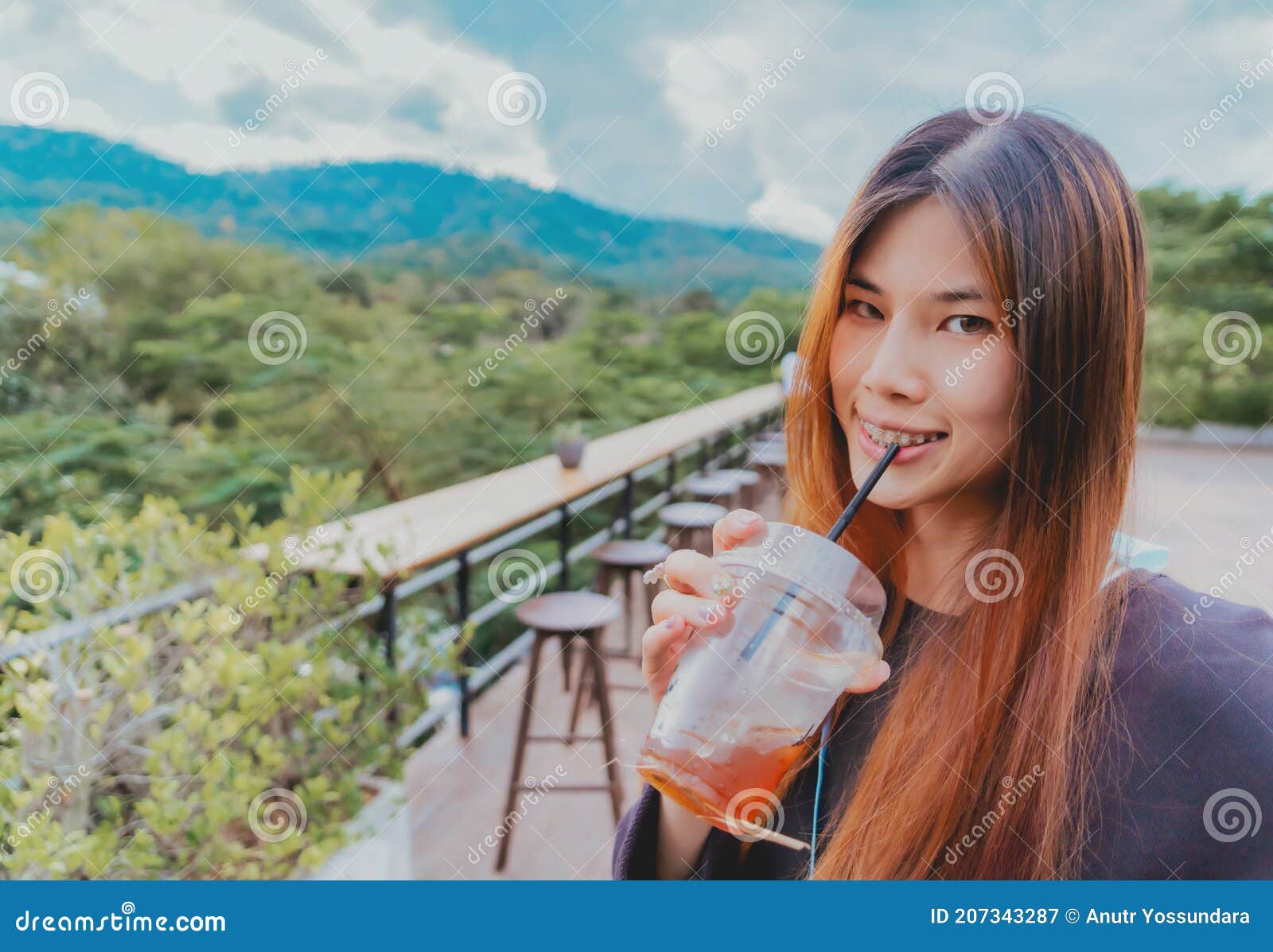 beauty smiling female is in cafe with forest and mountain nature background while drinking iced americano coffee