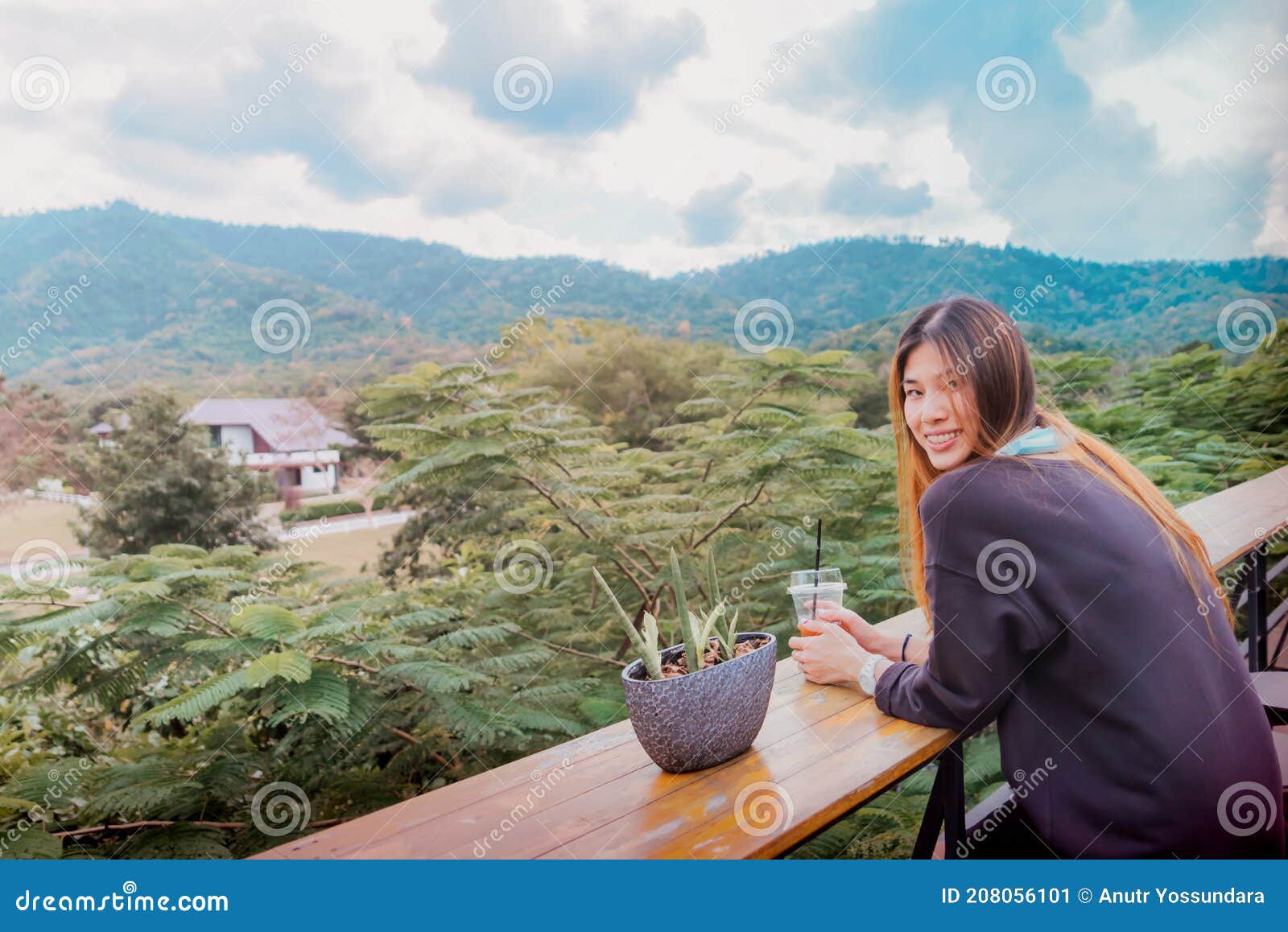 beauty young smiling female is sitting in cafe with forest and mountain nature background while drinking iced americano