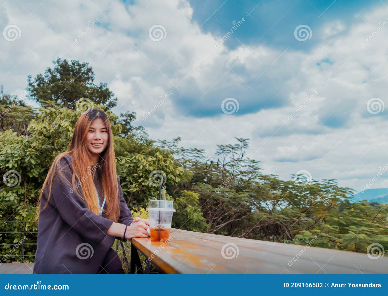 beauty young smiling female is sitting in cafe with forest and mountain nature background while drinking iced americano