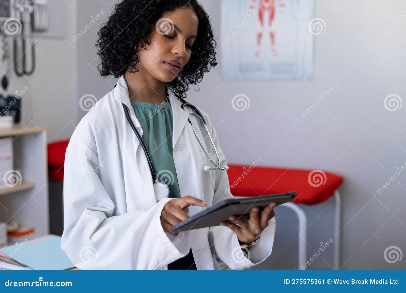 biracial female doctor wearing sthethoscope, using tablet at doctor's office