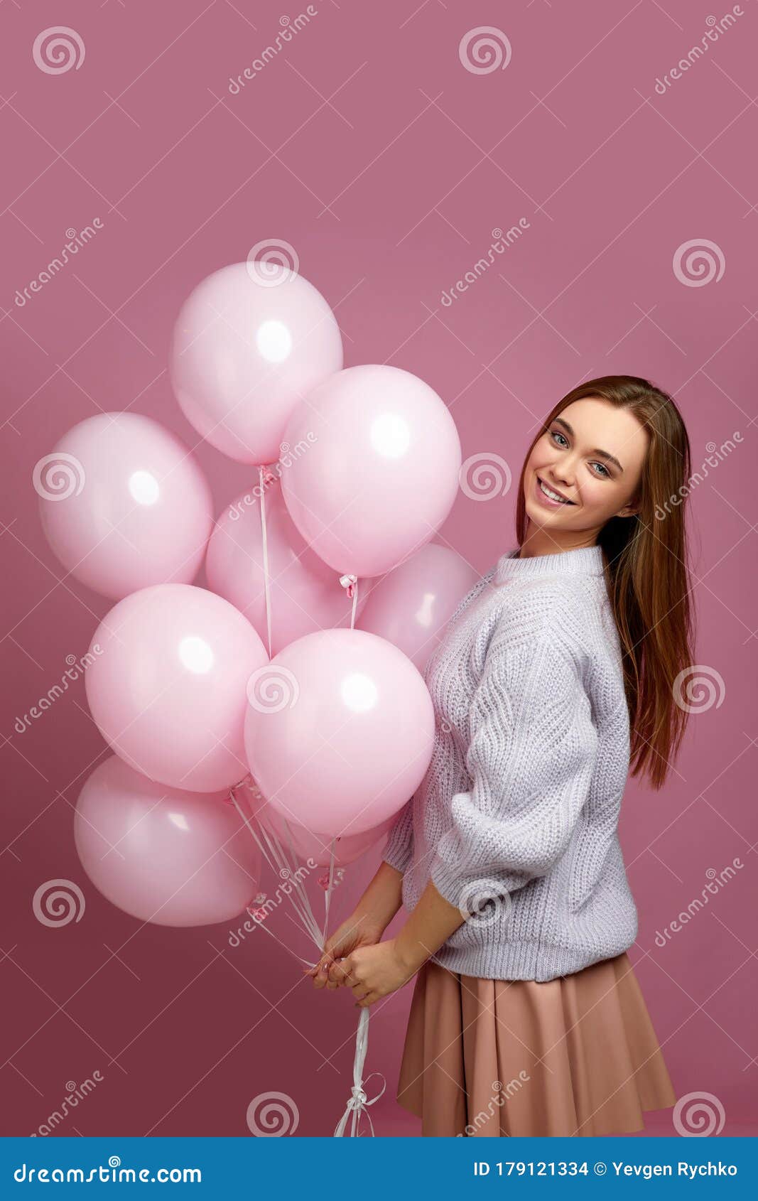 Cheerful birthday woman dressed in slumber suit smiles broadly has cream  around mouth holds glass of cocktail and doughnuts with candles poses near  table full of desserts surrounded by balloons - Stock