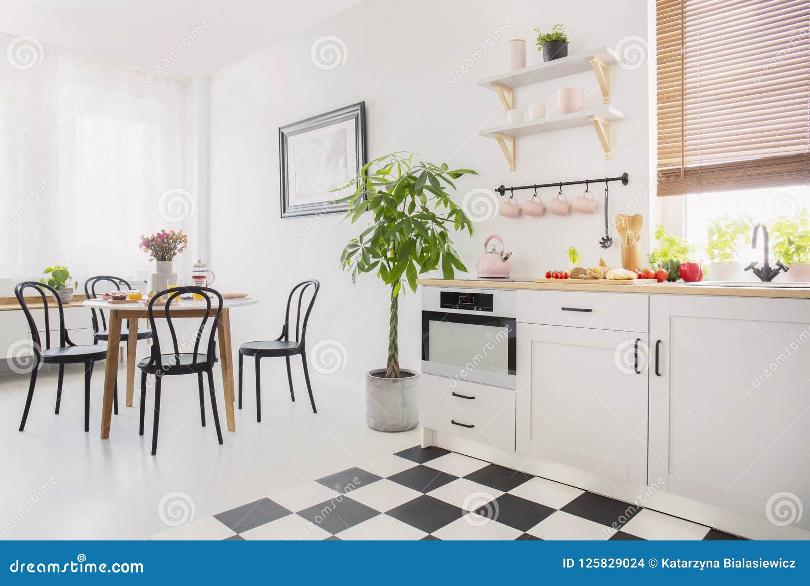 black chairs at dining table with flowers in white flat interior with plant next to kitchenette. real photo