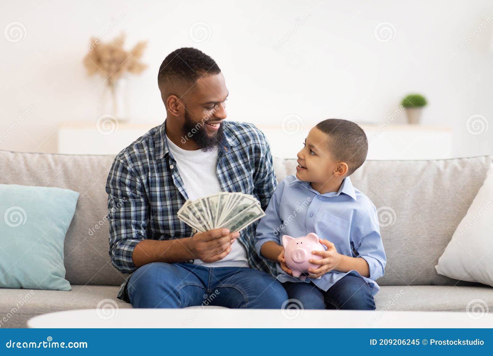 Black Father and His Son Putting Money in Piggybank Indoors Stock Image